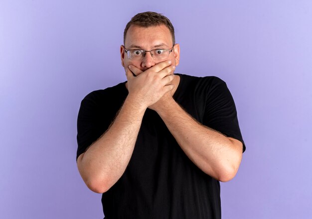 Overweight man in glasses wearing black t-shirt covering mouth with hands being shocked over blue