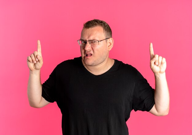 검은 색 티셔츠를 입은 안경에 과체중 남자가 분홍색 벽 위에 서서 검지 손가락을 보여주는 불쾌감