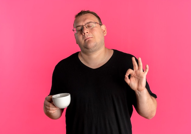 Overweight man in glasses wearing black t-shirt holding coffee cup doing ok sign pleased standing over pink wall