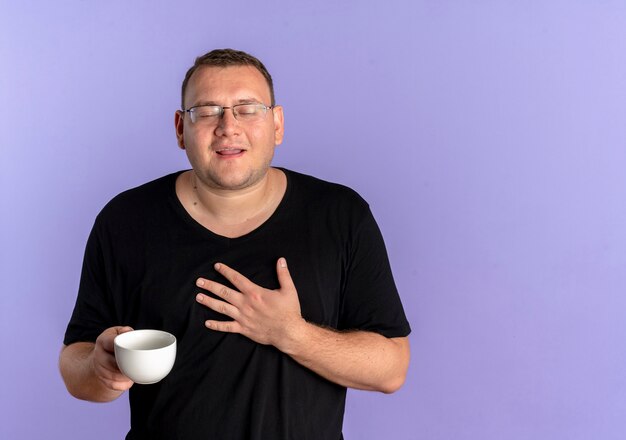 Полный мужчина в очках в черной футболке держит чашку кофе, держа руку на груди, чувствуя благодарность, стоя над синей стеной