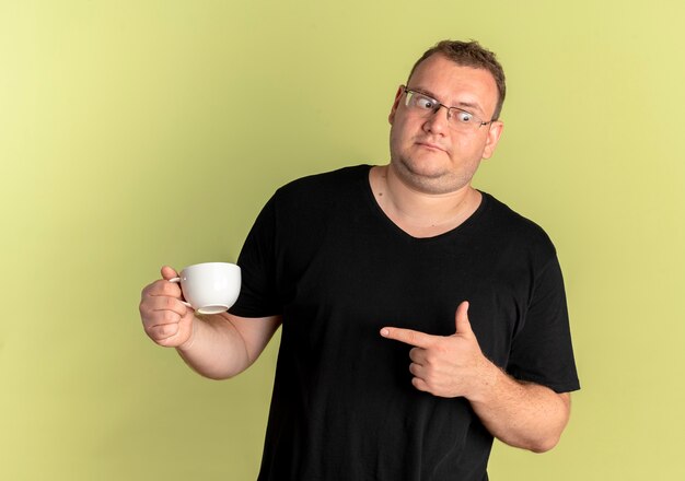 黒いTシャツを着た太りすぎの男性がコーヒーカップを持って指でそれを指さし、明るい壁の上に立って混乱している