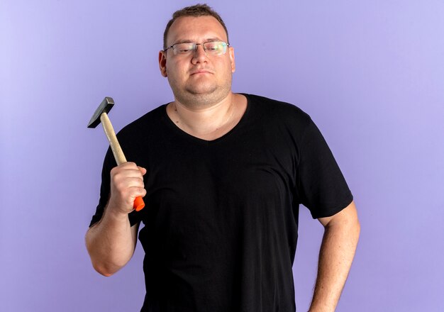 Overweight man in glasses wearing black t-shirt holding hammer looking confident over blue