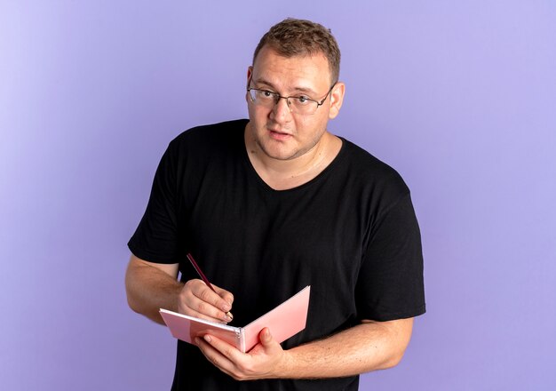 Overweight man in glasses wearing black t-shirt holding notebook writing something with serious face over blue