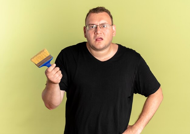 Overweight man in glasses wearing black t-shirt holding paint brush  as asking or arguing standing over light wall