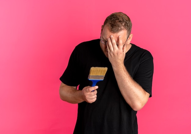 Foto gratuita uomo in sovrappeso con gli occhiali che indossa la maglietta nera che tiene il pennello che copre il viso con la mano delusa in piedi sopra il muro rosa