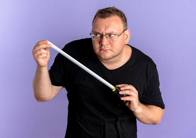 Overweight man in glasses wearing black t-shirt holding ruler looking aside like a spy standing over blue wall