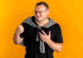 Free photo overweight man in glasses wearing black t-shirt holding smartphone clenching fist happy and excited standing over orange wall