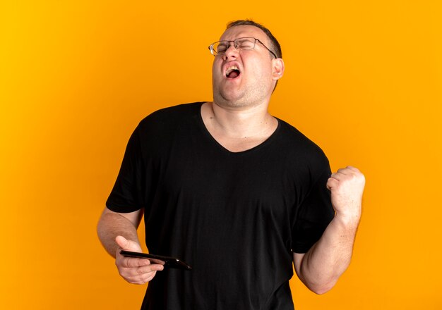 Overweight man in glasses wearing black t-shirt holding smartphone clenching fist shouting with aggressive expression standing over orange wall