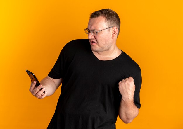 Overweight man in glasses wearing black t-shirt holding smartphone clenching fist with aggressive expression over orange