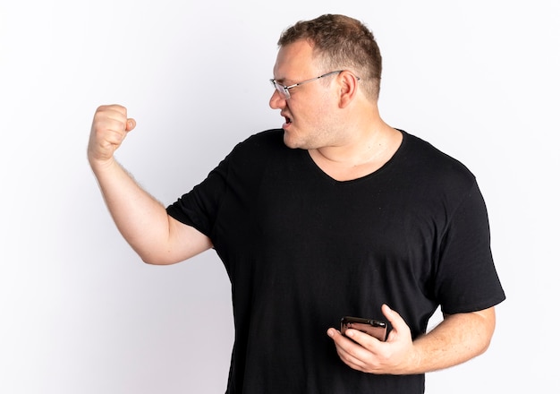 Free photo overweight man in glasses wearing black t-shirt holding smartphone clenching fist with angry face standing over white wall