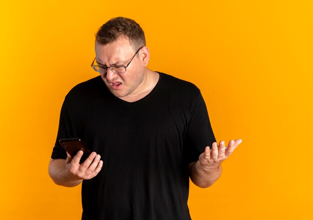 オレンジ色の壁の上に立っている混乱した表情で画面を見ているスマートフォンを保持している黒いTシャツを着て眼鏡をかけた太りすぎの男