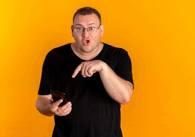 Overweight man in glasses wearing black t-shirt holding smartphone pointing with finger at it confused standing over orange wall