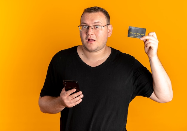 Полный мужчина в очках, одетый в черную футболку, держит смартфон, демонстрирующий внешний вид кредитной карты, удивлен оранжевым