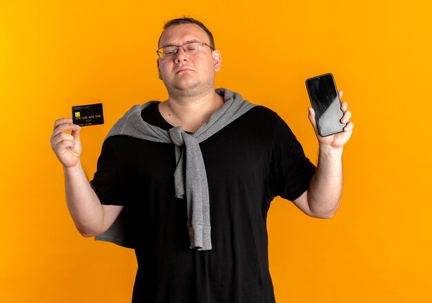 Overweight man in glasses wearing black t-shirt holding smartphone showing credit card looking confident over orange
