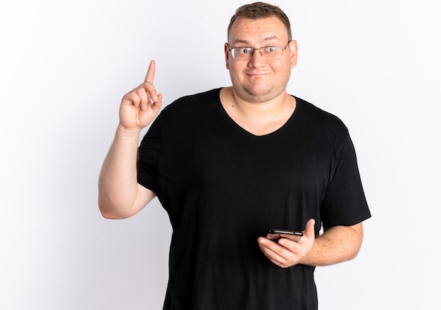 白い壁の上に立っている新しいアイデアを持っている人差し指を示すスマートフォンを保持している黒いTシャツを着た眼鏡の太りすぎの男