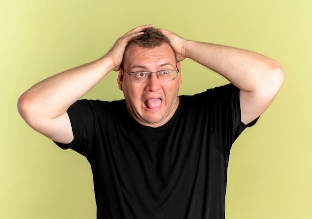 Overweight man in glasses wearing black t-shirt lookign aside pulling his hair in panic over light