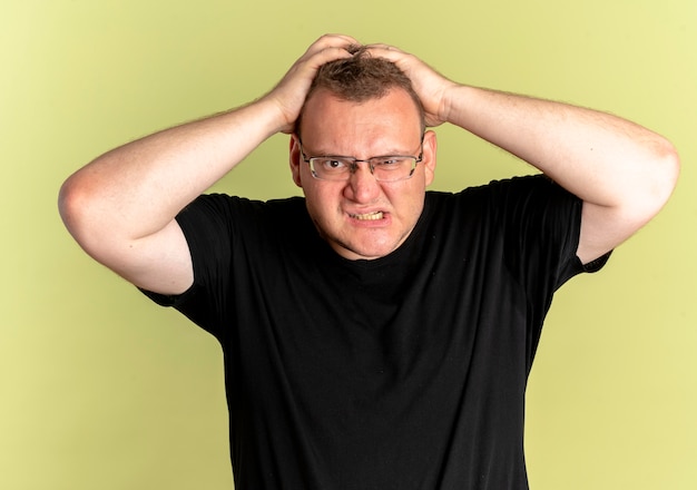 Overweight man in glasses wearing black t-shirt lookign at camera with angry face going wild pulling his hair over light