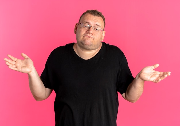 Foto gratuita uomo in sovrappeso con gli occhiali che indossa una maglietta nera che sembra confuso allargando le braccia ai lati senza risposta in piedi sul muro rosa