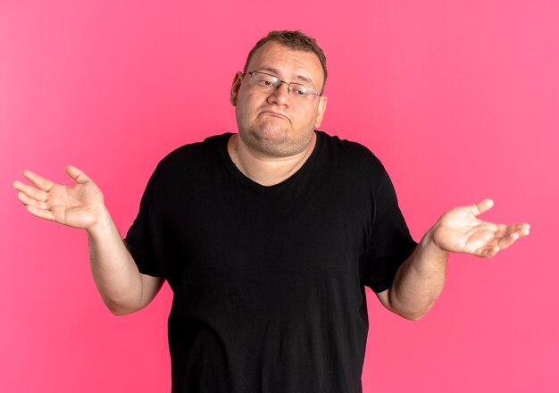 Overweight man in glasses wearing black t-shirt lookign confused spreading arms to the sides having no answer standing over pink wall