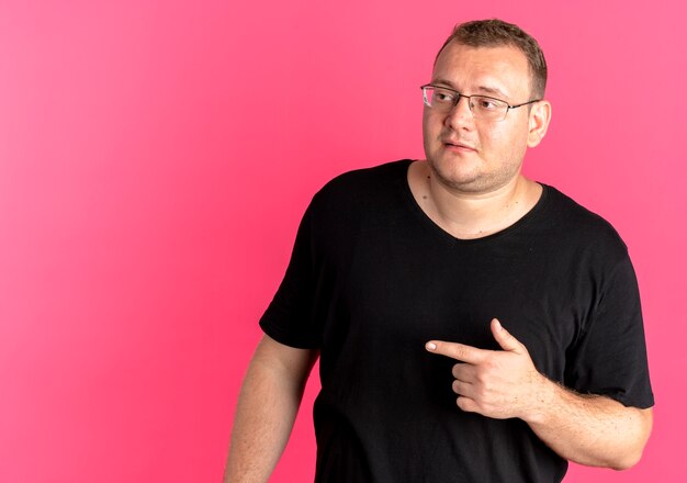 분홍색 벽 위에 서있는 측면에 검지 손가락으로 옆으로 보이는 검은 색 티셔츠를 입고 안경에 과체중 남자