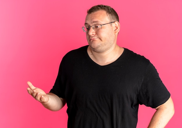 Overweight man in glasses wearing black t-shirt looking aside with arm out thinking or asking standing over pink wall