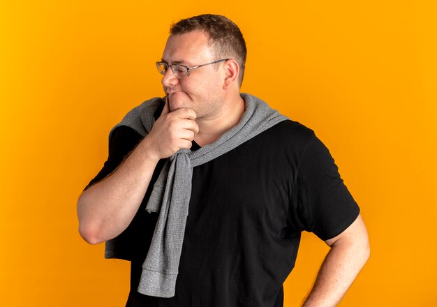 Overweight man in glasses wearing black t-shirt looking aside with hand on chin with pensive expression standing over orange wall