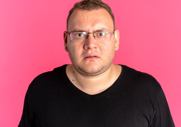 Overweight man in glasses wearing black t-shirt looking at camera surprised and displeased standing over pink wall