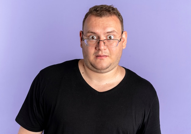 Free photo overweight man in glasses wearing black t-shirt looking at camera surprised standing over blue wall