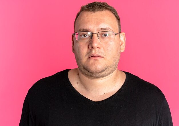Overweight man in glasses wearing black t-shirt looking at camera with sad expression standing over pink wall