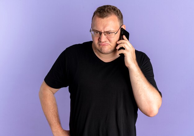 Overweight man in glasses wearing black t-shirt looking confused and displeased while talking on mobile phone over blue