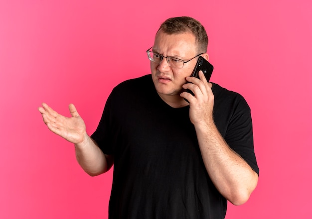ピンクで携帯電話で話している間、混乱して不快に見える黒いTシャツを着た眼鏡の太りすぎの男