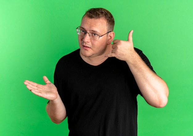 Uomo in sovrappeso con gli occhiali che indossa una maglietta nera che sembra confuso facendomi chiamare gesto sul verde