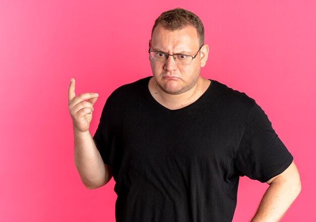 Overweight man in glasses wearing black t-shirt looking confused showing index finger thinking standing over pink wall