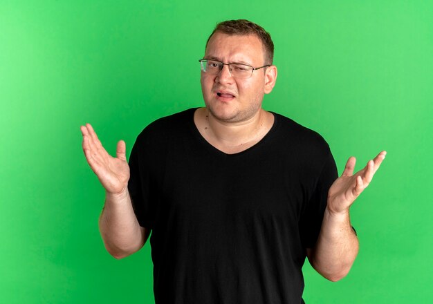 Overweight man in glasses wearing black t-shirt looking confused shrugging shoulders standing over green wall