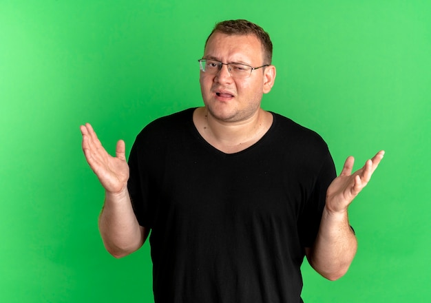 Полный мужчина в очках в черной футболке с растерянным видом пожимает плечами и стоит над зеленой стеной