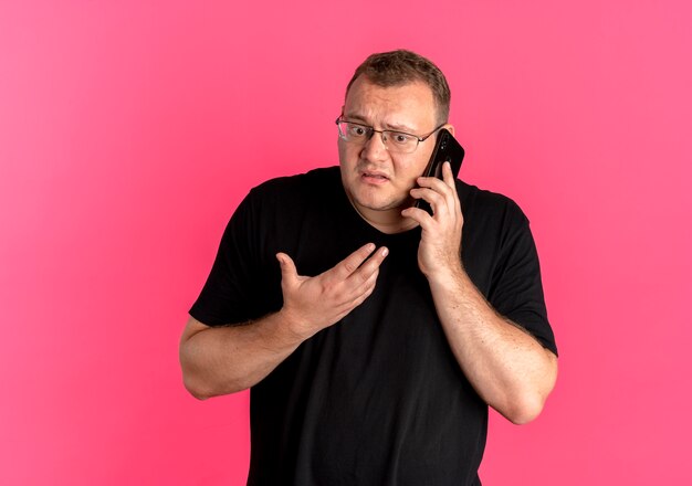 Overweight man in glasses wearing black t-shirt looking confused while talking on mobile phone standing over pink wall