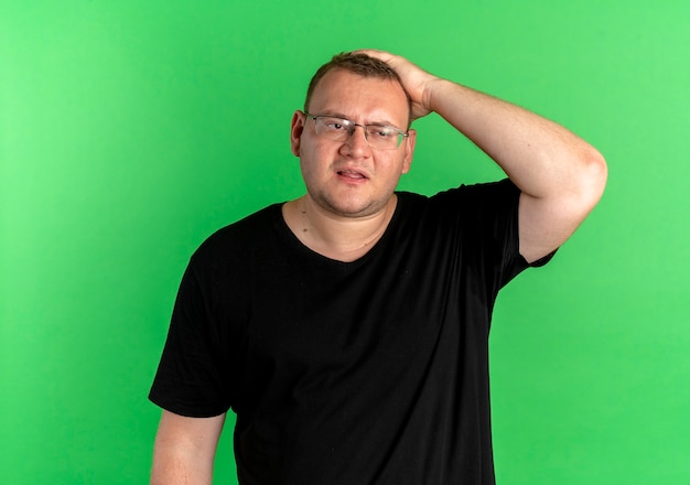 Free photo overweight man in glasses wearing black t-shirt looking confused with hand on his head for mistake forgot something important over green