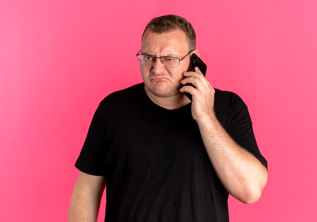 Overweight man in glasses wearing black t-shirt looking displeased while talking on mobile phone over pink