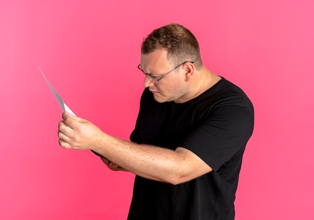 Overweight man in glasses wearing black t-shirt looking at document with serious face over pink