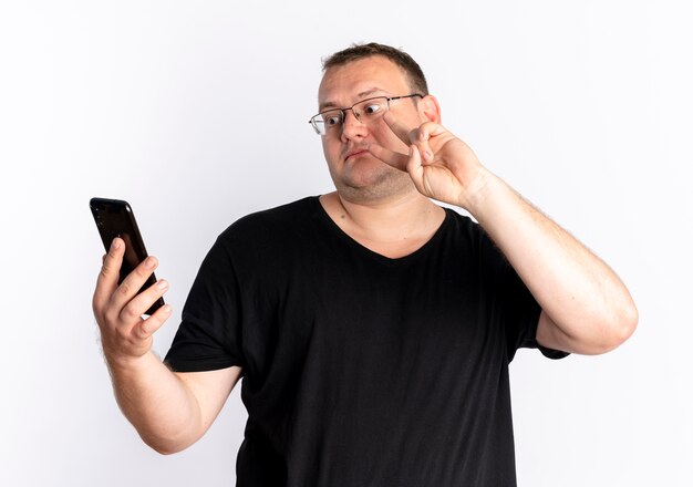 Overweight man in glasses wearing black t-shirt looking at his smartphone screen being online showing victory sign standing over white wall