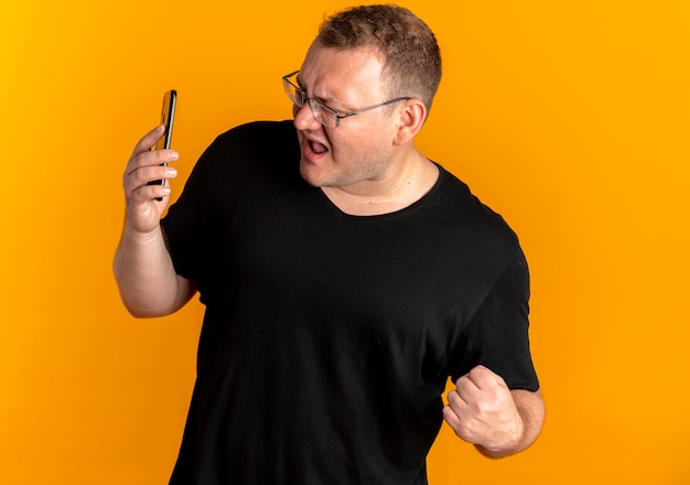 Overweight man in glasses wearing black t-shirt looking at his smartphone screen clenching fist very upset over orange