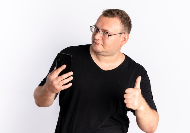 Overweight man in glasses wearing black t-shirt looking at his smartphone screen showing thumbs up standing over white wall