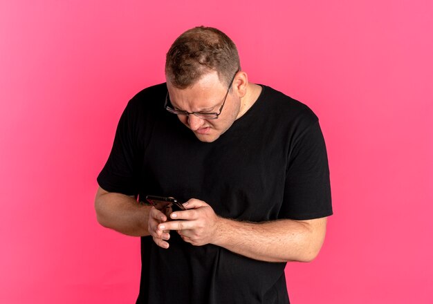 Overweight man in glasses wearing black t-shirt looking at his smartphone screen with angry face over pink