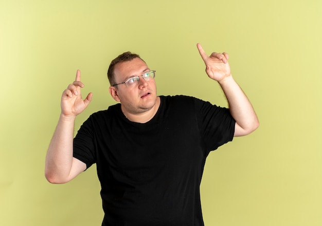 Overweight man in glasses wearing black t-shirt looking up confused pointing up with index fingers standing over light wall