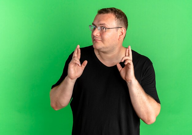 Overweight man in glasses wearing black t-shirt making desirable wish crossing fingers with happy face over green