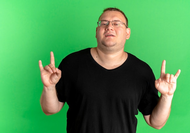 Free photo overweight man in glasses wearing black t-shirt making rock symbol looking confident over green