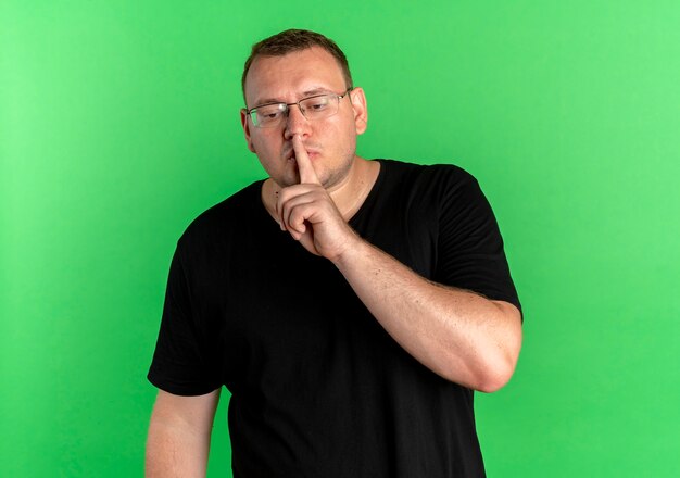 Overweight man in glasses wearing black t-shirt making silence gesture with finger on lips standing over green wall