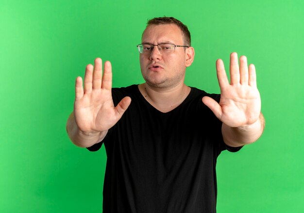 검은 티셔츠를 입고 안경에 과체중 남자는 녹색 벽 위에 서있는 손으로 노래를 중지합니다.