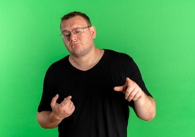 緑の壁の上に立っている人差し指で指している黒いTシャツを着て眼鏡をかけた太りすぎの男