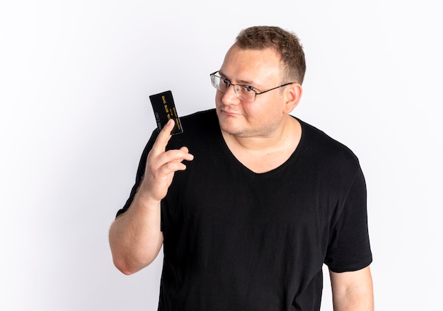 Overweight man in glasses wearing black t-shirt showing credit card looking at camera confused standing over white wall